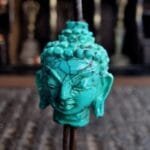 A hand-carved blue-green turquoise bead in the shape of a Buddha head, showing the detailed ushnisha and serene expression.