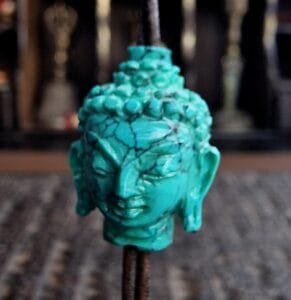 A hand-carved blue-green turquoise bead in the shape of a Buddha head, showing the detailed ushnisha and serene expression.