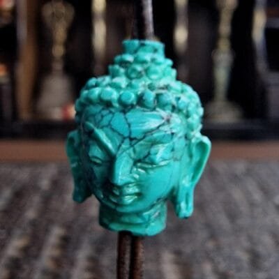 A hand-carved blue-green turquoise bead in the shape of a Buddha head, showing the detailed ushnisha and serene expression.